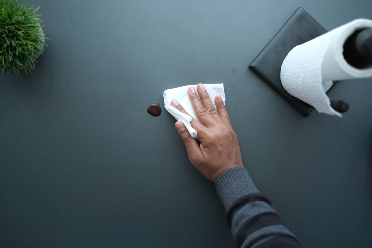 Une main essuie une surface sombre avec un mouchoir à proximité d’un rouleau de papier et d’une plante.